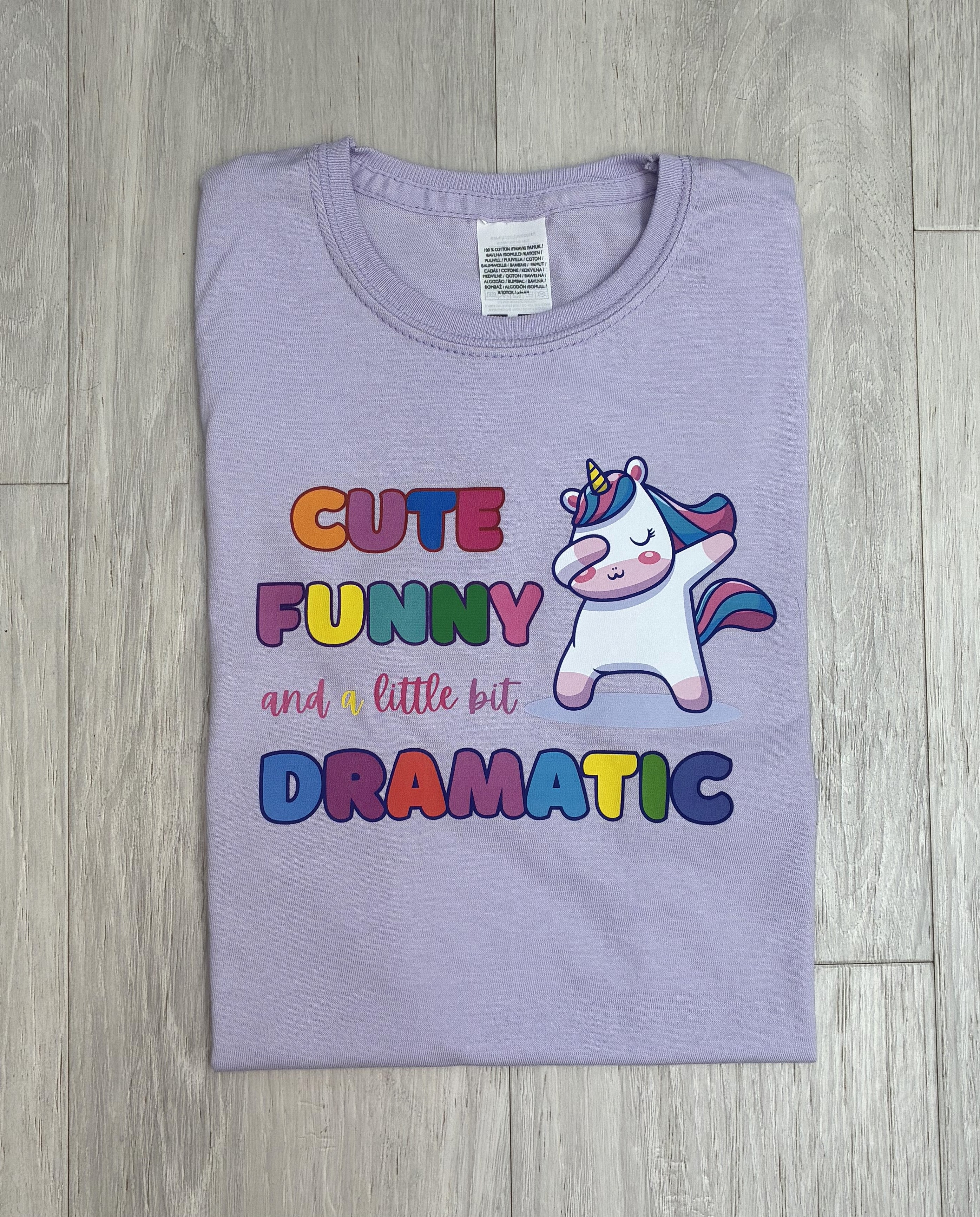 A folded liliac t-shirt with a colorful graphic print featuring the text 'CUTE FUNNY and a little bit DRAMATIC' in multicolored bubble letters next to a cartoon dancing unicorn, laid flat on a grey wood floor.