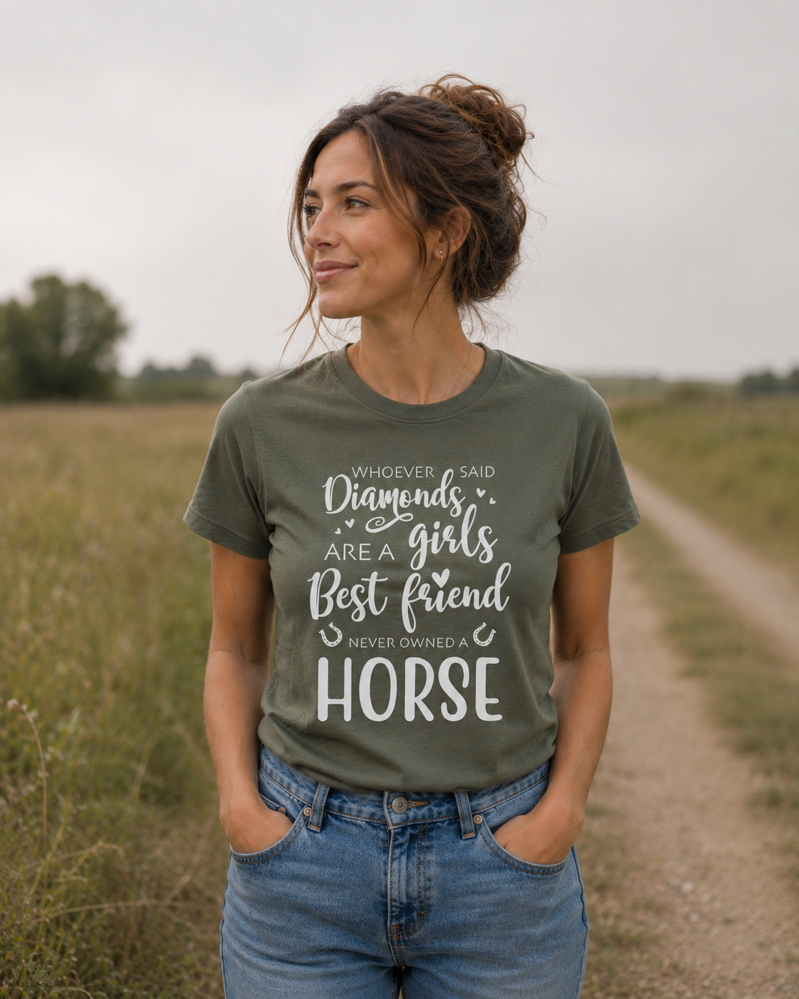 A woman standing outdoors on a dirt path wearing an olive green graphic t-shirt with white text that reads 'WHOEVER SAID Diamonds are a girls Best friend NEVER OWNED A HORSE'.