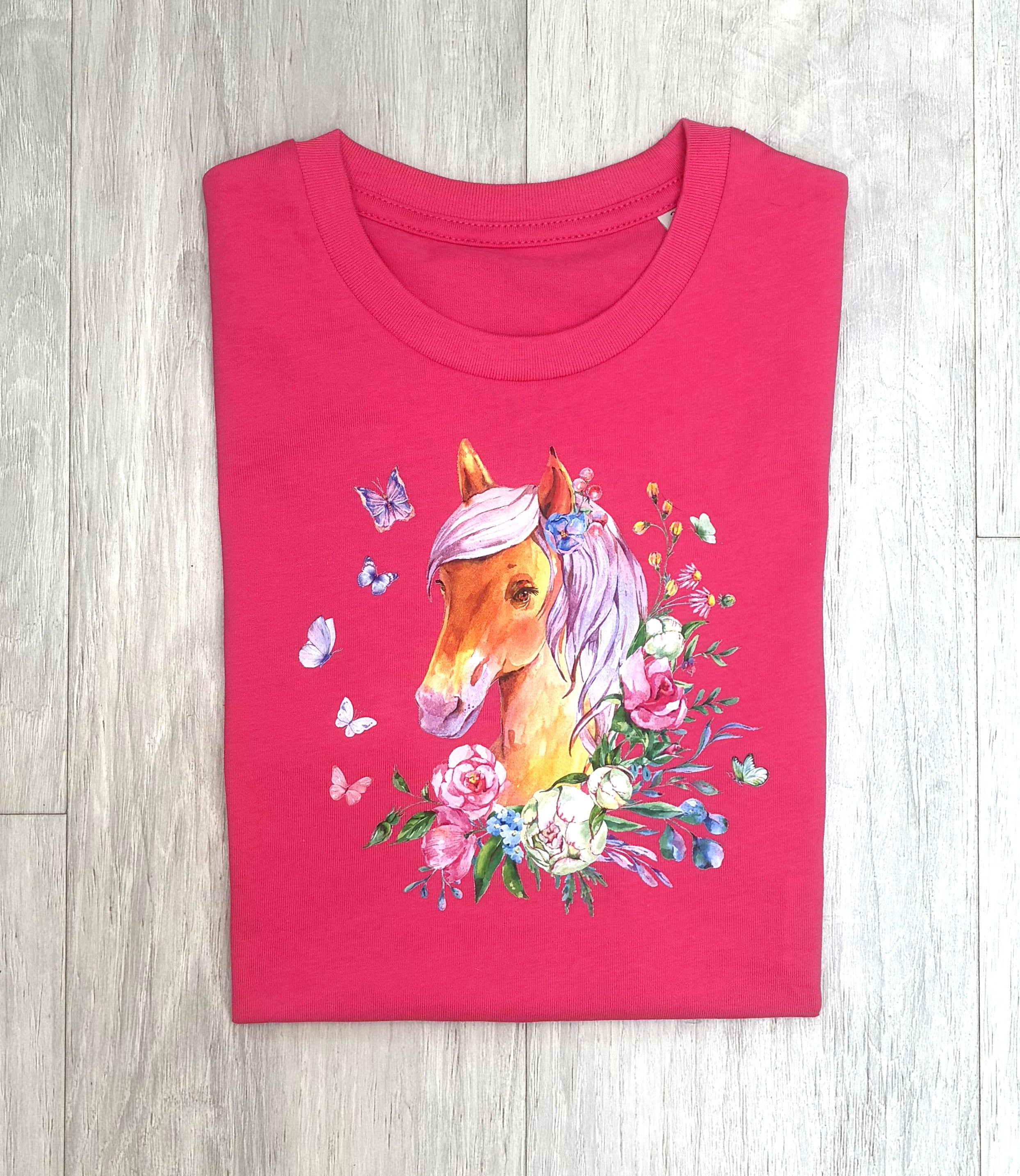 A folded hot pink t-shirt with a colorful graphic of a horse head surrounded by flowers and butterflies, laid flat on a light gray wood plank background.