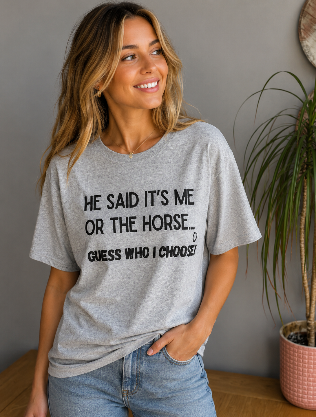 A woman wearing a light heather gray graphic t-shirt with black text that reads 'HE SAID IT'S ME OR THE HORSE.