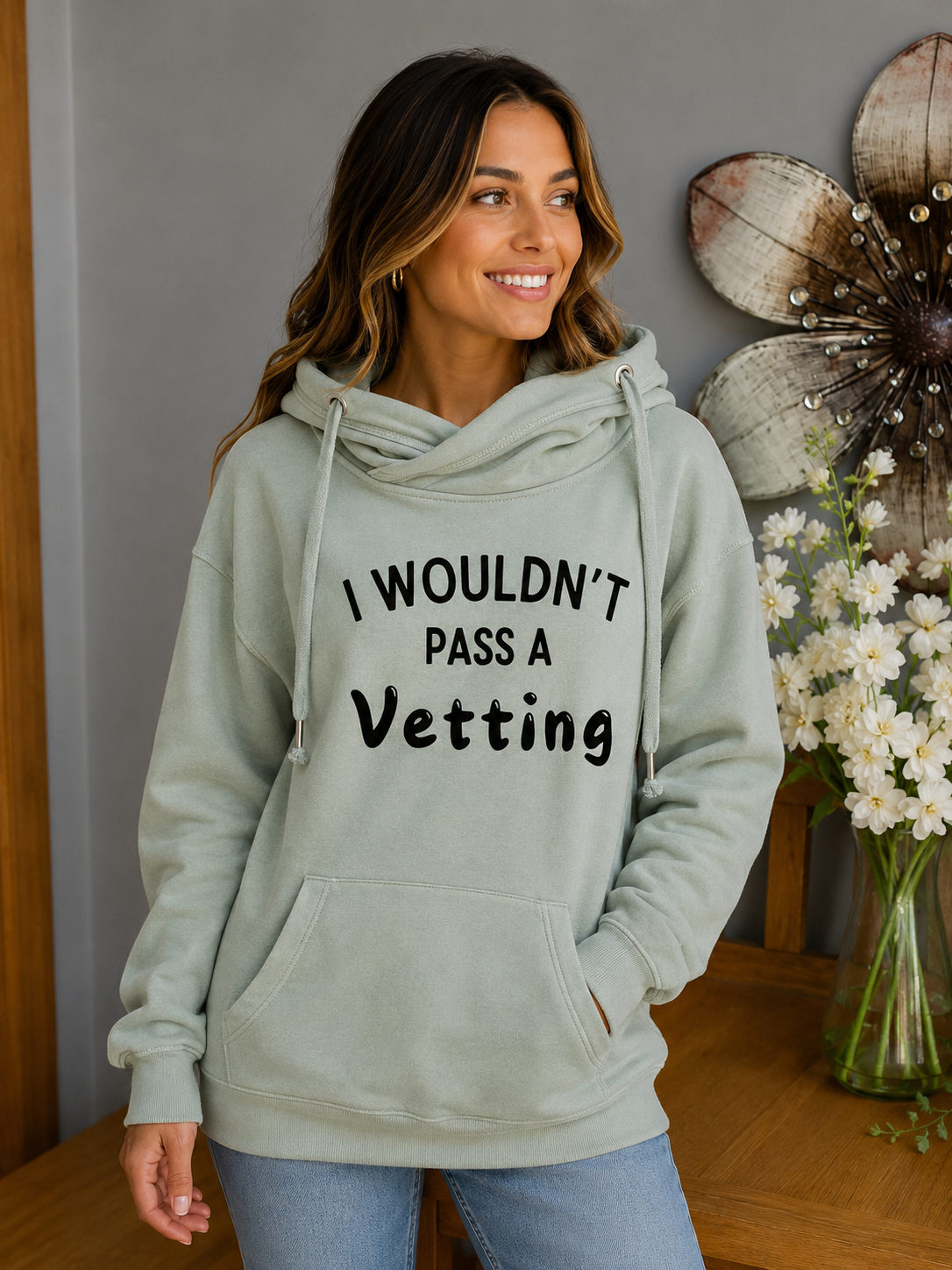 A woman wearing a sage green cowl neck pullover hoodie with the text 'I WOULDN'T PASS A Vetting' printed in black across the chest, paired with light blue jeans in an indoor setting.