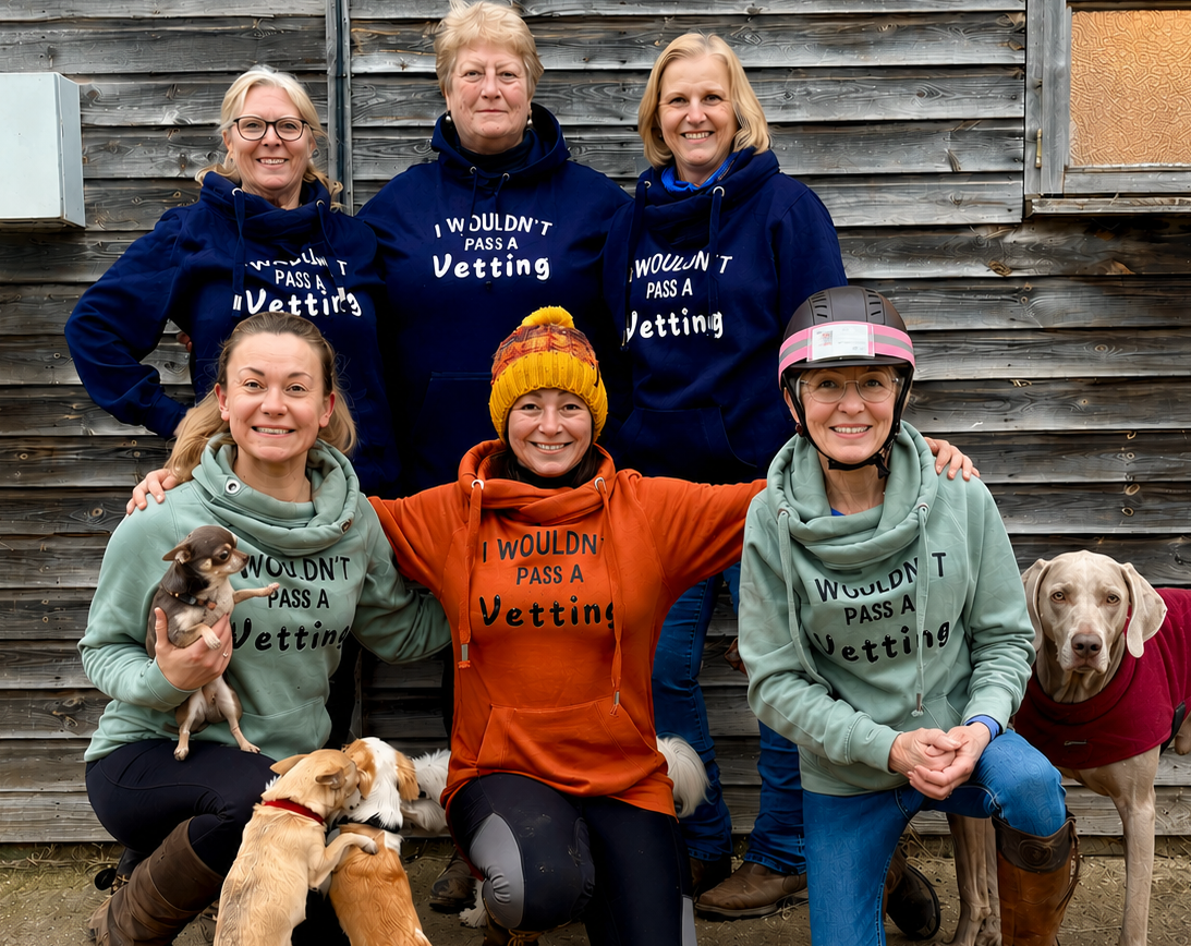Five women posing outdoors in front of a weathered wood building, wearing matching navy blue hoodies with white text that reads 'I WOULDN'T PAN A VetTErIN''. Two dogs are also present, one on the left being held and one standing on the right in a red sweater.