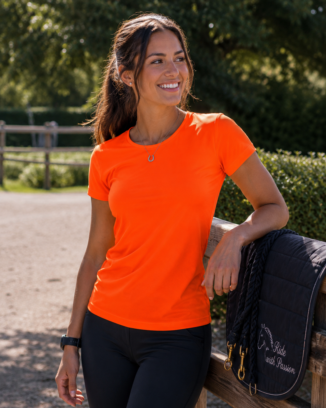 A woman wearing a bright orange short-sleeve athletic t-shirt and black riding pants leans against a wooden fence outdoors, with a black quilted saddle pad draped over the rail. Trees and a fence are visible in the background.
