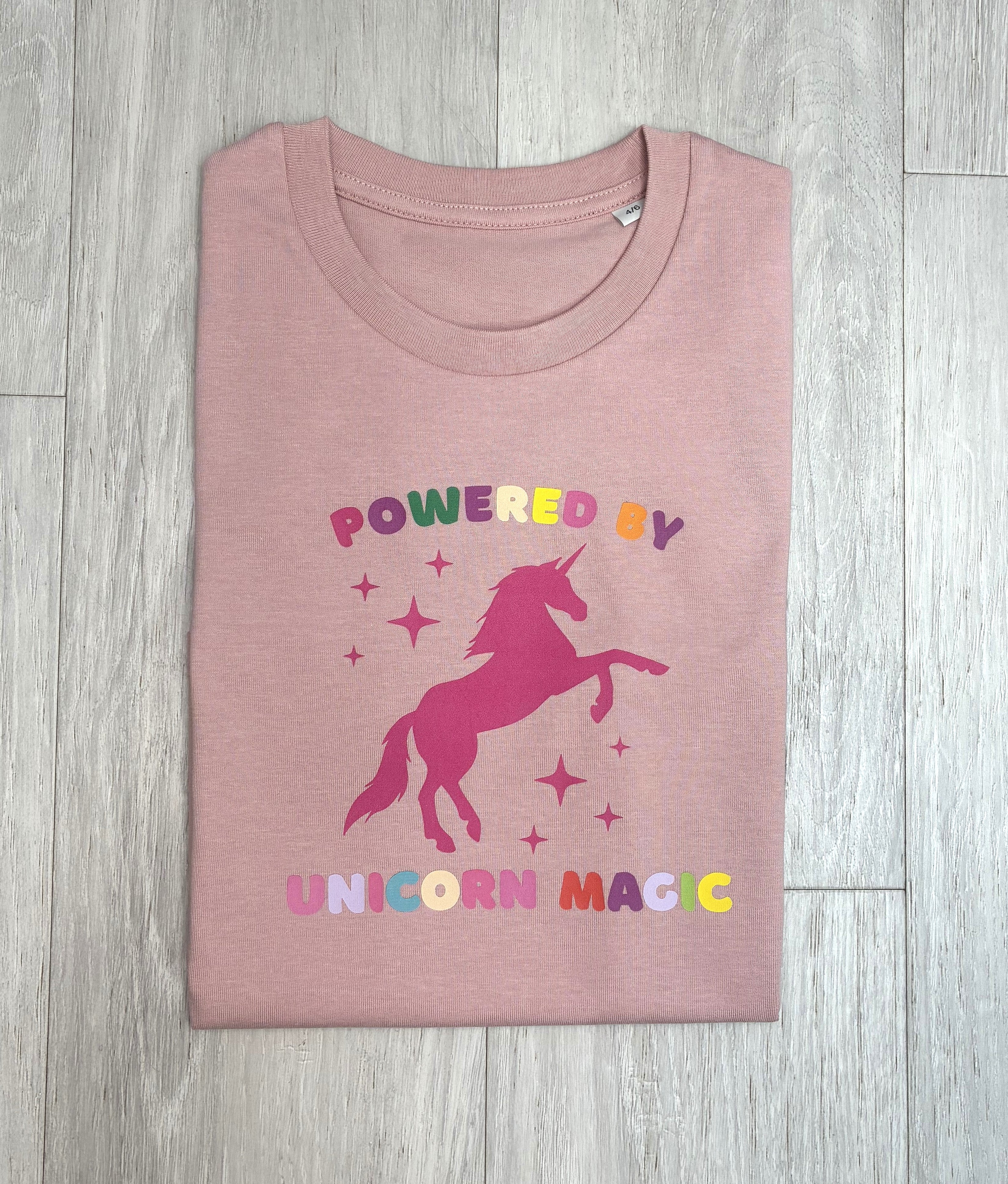 A folded dusty pink t-shirt with a colorful graphic of a pink unicorn and the text 'POWERED BY UNICORN MAGIC' in multicolored letters, laid flat on a light grey wood plank background.