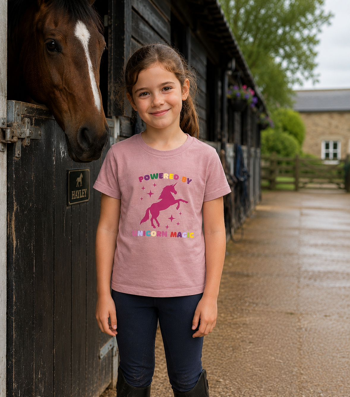 A young girl stands in front of horse stalls wearing a dusty pink graphic t-shirt with a pink unicorn and the words 'POWERED BY KNOWLEDGE'. A brown horse is visible in the stall next to her, and a wet stone path stretches into the background.