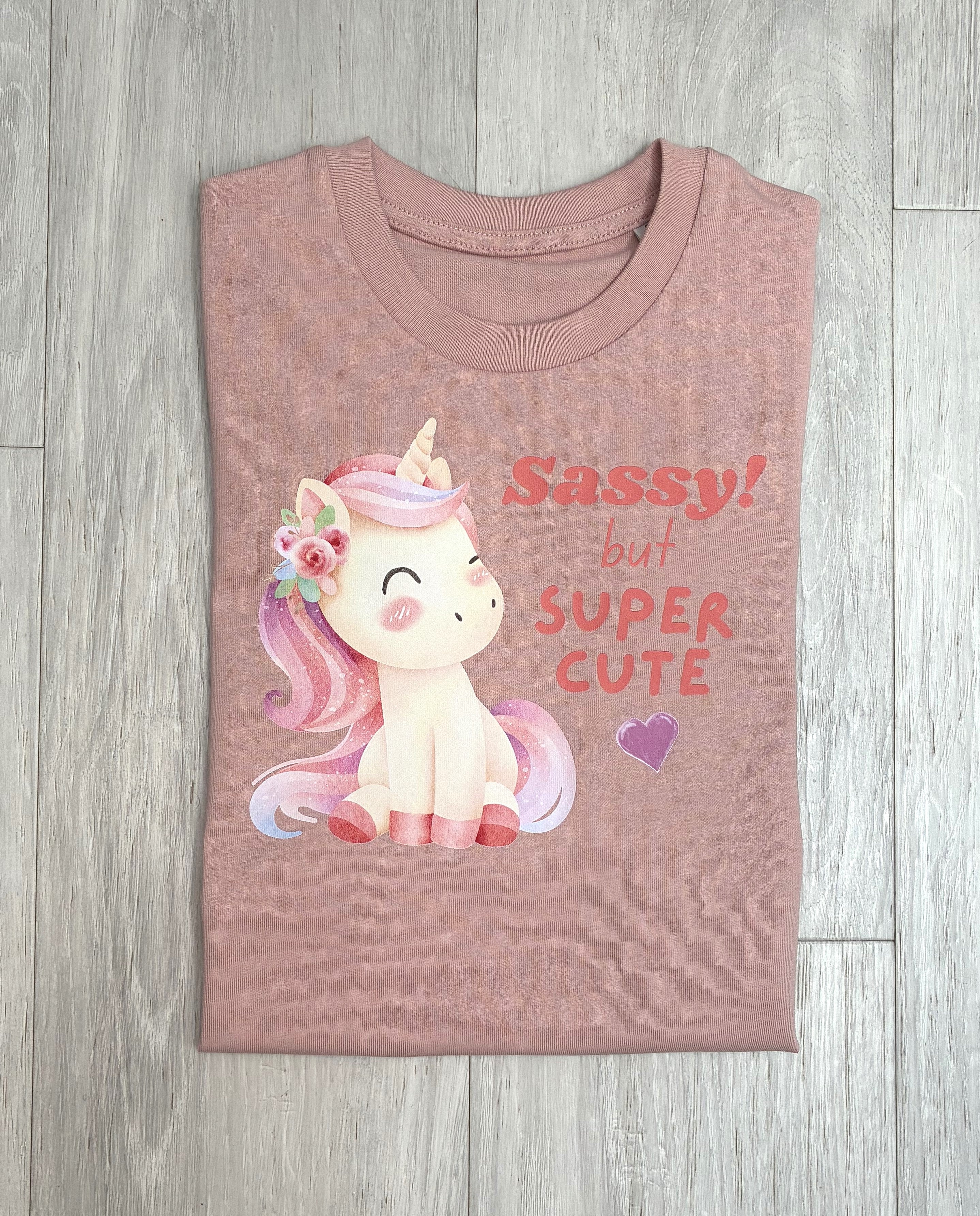 A folded dusty rose pink t-shirt with a cartoon unicorn graphic and the text 'Sassy! but SUPER CUTE' printed on the front, laid flat on a grey wood-plank floor.