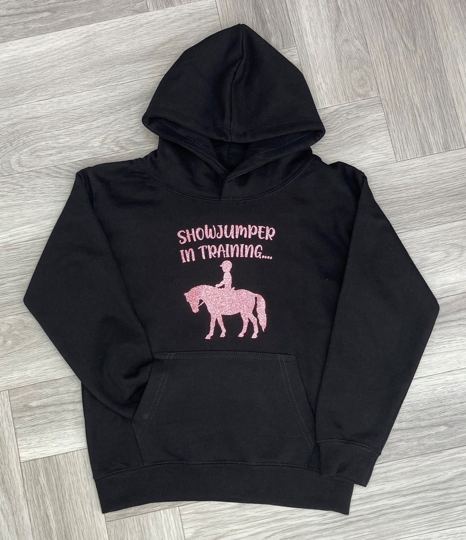 A black hoodie laid flat on a grey wood-look floor, featuring pink text that reads 'SHOWJUMPER IN TRAINING.