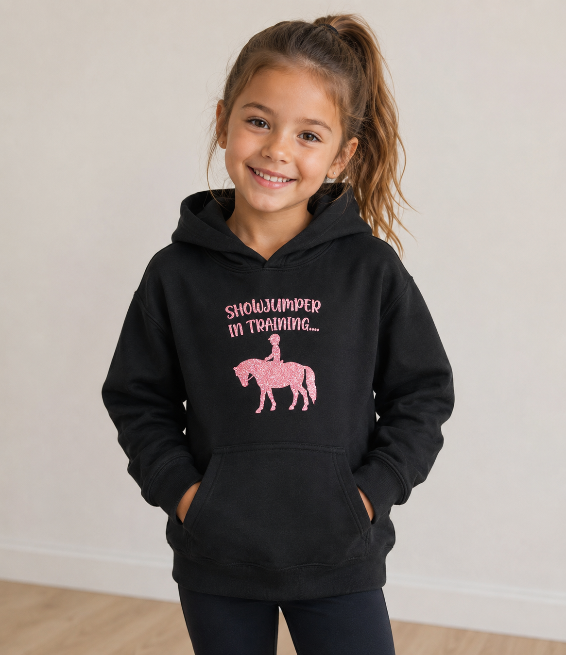A young girl with her hair in a ponytail smiles while wearing a black pullover hoodie with a pink graphic that reads 'SHOWJUMPER IN TRAINING.