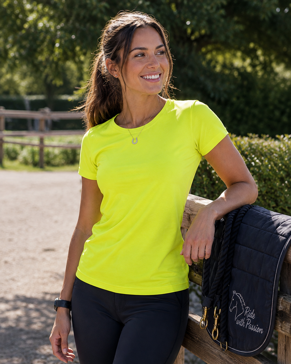 A woman wearing a bright neon yellow short-sleeve athletic t-shirt and black leggings leans against a wooden fence outdoors, with a dark navy equestrian saddle pad featuring the words 'Ride with Passion' draped over the fence.